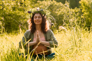 Woman meditating in tall grass wearing flower wreath under summer sunlight