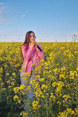 Fototapeta premium Aesthetic young woman enjoys the rapeseed field, smelling the beautiful yellow flowers. Girl in pink shirt adds charm to the picturesque scenery under the clear blue sky