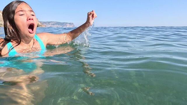 A woman in a turquoise bikini is submerged in the ocean, her face contorted in a scream, her right hand raised out of the water. Summer holiday vacation and travel concept
