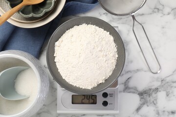 Digital kitchen scale with bowl of flour and other products on white marble table, flat lay