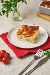 Portion of lasagne with salmon and broccoli on a light plate on a light concrete background.