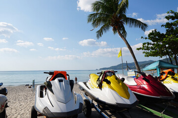 Jet skis lined up on a sandy beach with palm trees and calm ocean under bright sunny sky