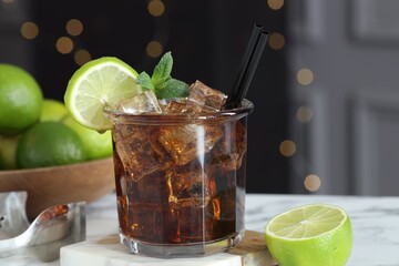 Tasty Cuba Libre cocktail in glass, limes and tongs on white marble table, closeup. Bokeh effect
