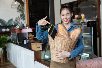 Asian woman holding basket filled with fresh baguette bread standing in small bakery cafe, smiling and pointing at promotion sign. Denim shirt and apron showing cheerful small business concept.