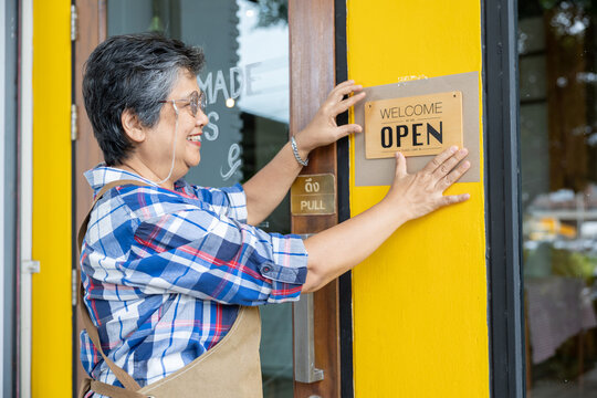Asian senior woman smiling while placing open welcome sign on storefront yellow wall. Wearing plaid shirt with apron representing confident entrepreneur running local small business shop. - Powered by Adobe