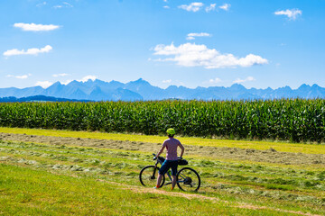 Woman cyclist standing on green meadow in spring season with Tatra Mountains peaks in background, Poland