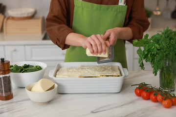 Woman grating cheese onto spinach lasagna at marble table indoors, closeup