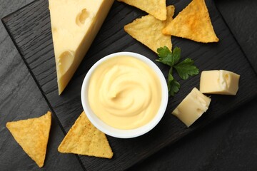 Tasty dipping cheese sauce in bowl, nacho chips and parsley on black table, top view