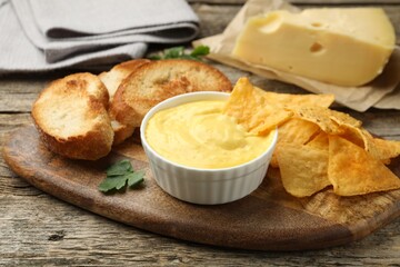 Tasty cheese dipping sauce in bowl, nacho chips and croutons on wooden table, closeup