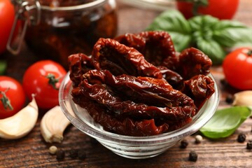 Delicious sun dried tomatoes in glass bowl, fresh vegetables and spices on wooden table, closeup