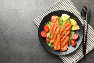 Tasty grilled salmon served with cutlery, lettuce, tomatoes and lemon on grey table, top view. Space for text