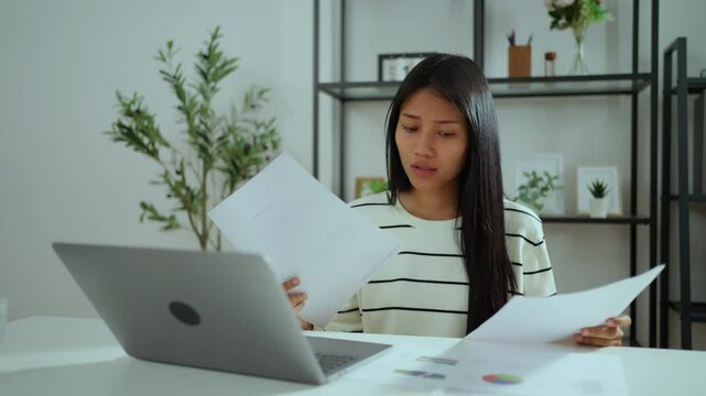 Young asian woman sitting at desk checking paperwork while working remotely, focused and serious expression, analyzing financial data or handling business report in modern home office setting.