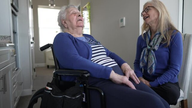 A woman in her 90s sits in a wheelchair, smiling as her daughter provides care and companionship during a visit at their cozy home together.