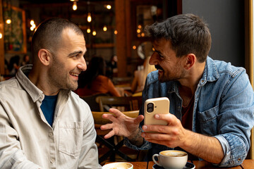 Two male friends enjoying their cappuccino at the cafe and showing interesting content on smartphone