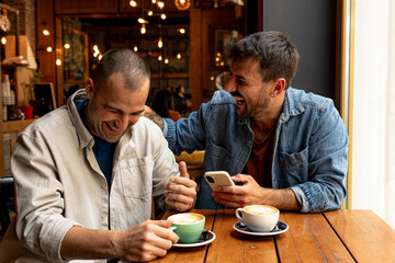 Two male friends are enjoying their time together in a cafe, laughing and showing something funny on smartphone