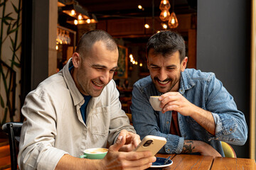 Two male friends enjoying coffee together, laughing and sharing funny content on a smartphone in a trendy cafe setting