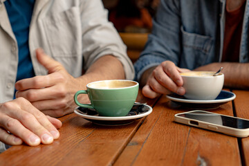 Close-up of two people holding cups of cappuccino at a cafe, talking and enjoying their coffee break, with a smartphone on the wooden table