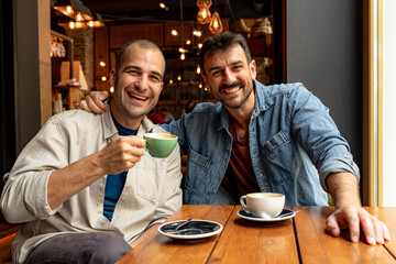 Two male friends enjoying coffee together, sharing smiles and laughter in a cozy cafe, creating a warm atmosphere of friendship