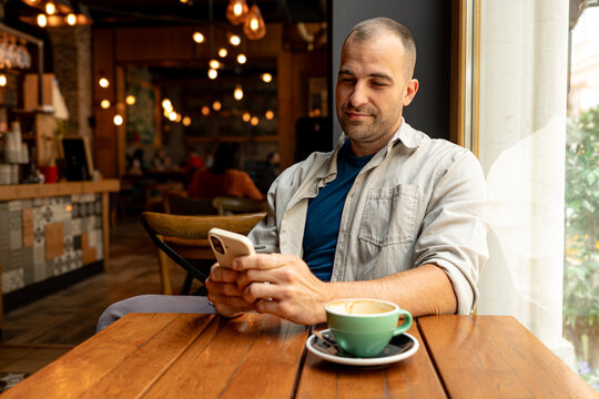 Man sipping cappuccino while browsing social media on a smartphone, enjoying a relaxed morning in a cozy cafe setting