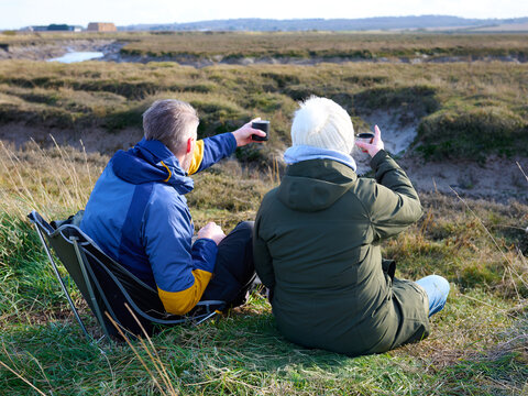 A senior woman and her adult son sit and relax with a hot drink in a grassy, marsh landscape, they are pointing at the view. - Powered by Adobe