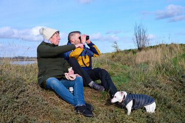A senior woman and her adult son sit and relax with a binoculars and a snack in a grassy, marsh landscape with their dog on a partly cloudy day.