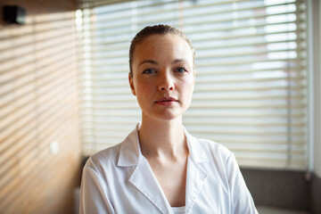 Confident female doctor in lab coat standing with arms crossed in medical office
