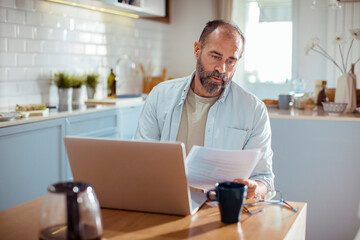 Mature man reviewing documents and working on laptop at kitchen table