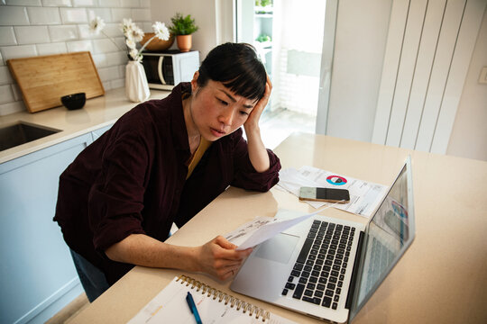 Stressed woman reviewing bills and finances on laptop at kitchen table