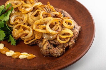 Grilled pork steak with golden fried onions served on a rustic clay plate. Traditional Eastern European dish with mustard, mayonnaise and fresh parsley garnish, on a light background