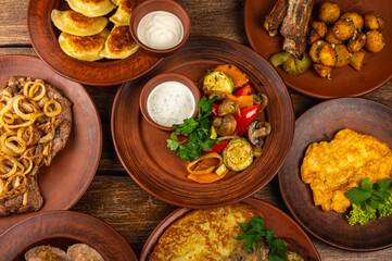 Traditional Eastern European cuisine served on rustic clay plates. Variety of homemade dishes including meat, potatoes, pancakes, dumplings, and grilled vegetables, on a wooden background