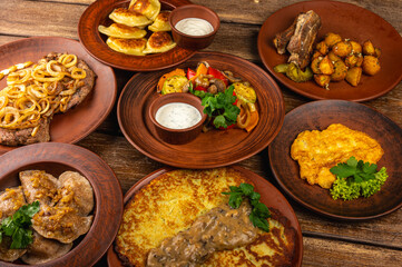 Traditional Eastern European cuisine served on rustic clay plates. A variety of homemade dishes including meat, potatoes, pancakes, dumplings and grilled vegetables, on a rustic background, top view.