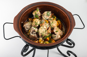 Traditional meat stew with vegetables served in a clay pot on a metal stand. Rustic presentation of hearty food, garnished with fresh herbs. Isolated on white background