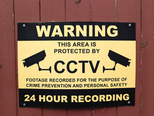 Yellow and black CCTV warning sign mounted on wooden fence detailing 24-hour surveillance for property security and crime prevention