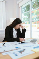 An overworked Asian businesswoman sits at her desk in a modern office, stressed and exhausted,...