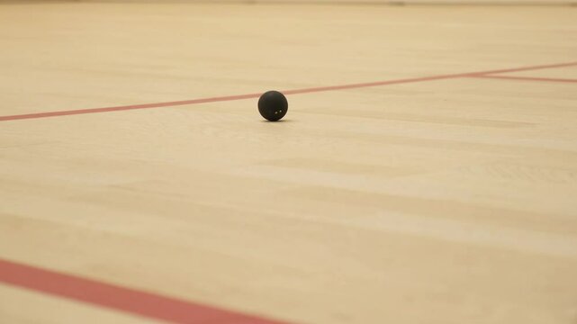 Squash ball captured mid-roll on the wooden floor of a squash court. The ball, visible with distinct yellow dots, showcases its circular motion against the court's markings
