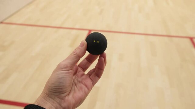 Close-up of a hand holding a black squash ball with two yellow dots, set against a wooden squash court with red boundary lines. The scene suggests readiness for a game