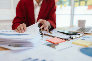 A confident young Asian businesswoman sits at her desk with a laptop and documents, smiling while managing office tasks and communication.