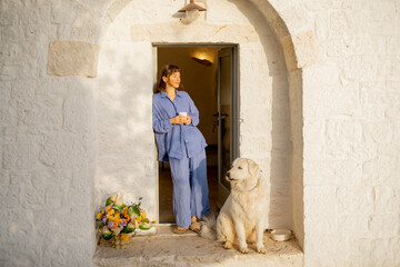 A woman in soft linen pajamas enjoys morning coffee beside her fluffy white dog, standing in the...