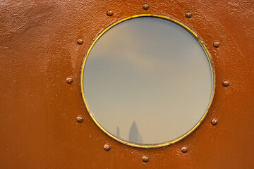 A detailed closeup of a round porthole with a protective glass with soft sky reflection. Brown...