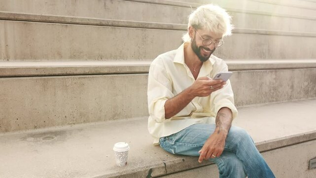 A young, stylish man with bleached blonde hair sits on steps, enjoying his coffee and scrolling on his phone. He smiles and laughs.