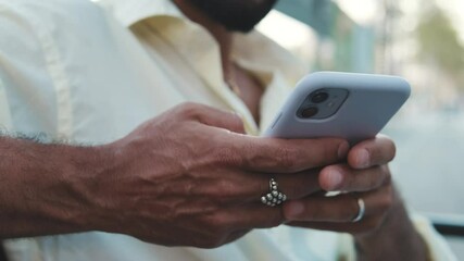 A man using a smartphone while commuting. He is wearing a white shirt and has rings on his fingers. The background shows city life out of a bus window. - Powered by Adobe