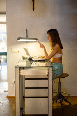 A woman working on her laptop at a modern glass desk in a sunlit home. The quiet morning light and...