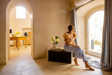 A woman sits barefoot by a sunlit door in a traditional Matera home. This image radiates calm,...