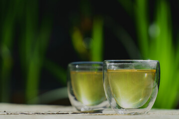 Two small transparent glass cups of green herbal tea placed on natural wooden surface outdoors ona sunny morning