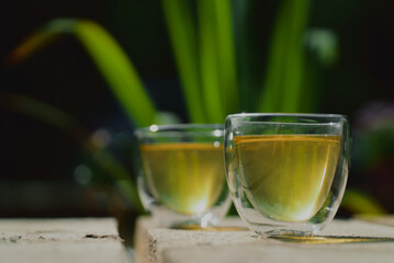 Two small transparent glass cups of green herbal tea placed on natural wooden surface outdoors ona sunny morning
