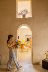 A woman arranges yellow flowers on a rustic wooden table in a stone kitchen. The scene reflects simplicity, tradition, and cozy Mediterranean living perfect for interior or lifestyle use