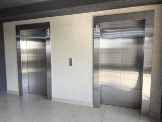 Closeup of two modern stainless steel elevator doors in light tiled hallway with textured wall, reflecting ambient light and emphasizing clean lines, symmetry and minimalism in architectural space