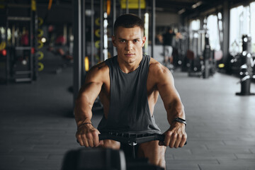 Focused handsome man using rowing equipment in modern gym. Fit young guy exercising on rowing machine at fitness center. Weight loss and cardio fitness concept