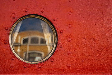A detailed closeup of a circular ship porthole set in a vivid red metal wall, reflecting the yellow and white facade of the historic customs office in Tervasaari harbor, Hamina, Finland.

