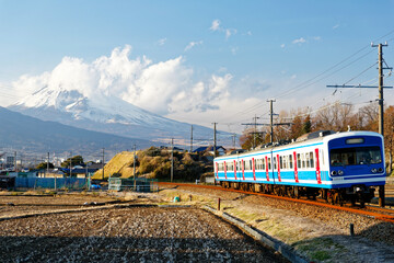 Obraz premium A local train of Izu-Hakone Railway traveling through harvested farmlands with the snow capped mountaintop of Fujisan veiled by clouds in background on a sunny winter day, in Mishima, Shizuoka, Japan